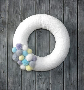 An image of a white round wreath hanging on a grey wood door. The white section is knitted in a chenille yarn. On the bottom left hand side is a collection of different size knitted balls and fluffy pom poms in mint, grey, yellow and white