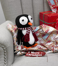 Load image into Gallery viewer, A penguin knitted toy sitting on some wrapping paper on an armchair. Knitted in black and white, with an orange beak and feet. The eyes are large white circles with black buttons sewn in the centre. Wearing a wine and white striped scarf
