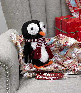 A penguin knitted toy sitting on some wrapping paper on an armchair. Knitted in black and white, with an orange beak and feet. The eyes are large white circles with black buttons sewn in the centre. Wearing a wine and white striped scarf