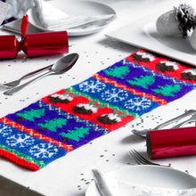 Load image into Gallery viewer, A festive table runner shown on a white table cloth. The runner is knitted in strips - red with 3 Christmas puddings, royal blue with 3 green Xmas trees and blue with 3 white snowflakes. The strips are separated by a thin band with zig zag detail
