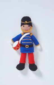 Drummer boy toy wearing black boots, red trousers, royal blue jacket and tall black hat. The jacket has gold, shoulder and button trims, the hat has gold details and chin strap. He carries a red and white drum on a white strap