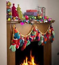 Load image into Gallery viewer, Advent Garland hanging on a mantlepiece decorated with Christmas ornaments and gifts. The garland has a yellow cord and alternating mitts and mini stockings with numbers embroidered. Knitted in bright yarns with stripes, contrast bands, and spots
