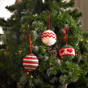 3 Christmas tree bauble ornaments on a tree. 1 is knitted in festive red, green and white stripes. 1 is white with two rows of red zig zags around the middle. 1 is green top and bottom with a white centre band with red hearts and edged with red