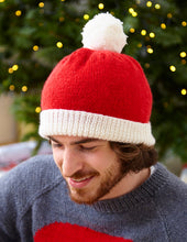 Load image into Gallery viewer, Man wearing a red and white beanie hat. The main section is knitted in stocking stitch with bright red DK yarn. The band is a white turn back knitted in rib. The hat is topped with a white pompom 
