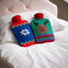 Load image into Gallery viewer, Two festive hot water bottle covers. 1 has a red band top and bottom. The middle royal blue panel has a white snowflake and is bordered by a green zig zag. 1 is green with purple, red and green stripes at the bottom, a red top and a reindeer head
