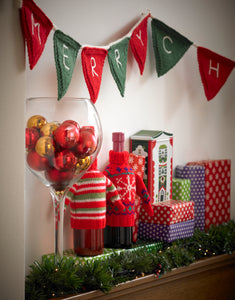 Image of a mantlepiece decorated for Christmas. Above is a traditional garland or bunting with green and red triangles. Each triangle has a letter spelling Merry Christmas. Also shown are snowflake motif and red, green and white wine bottle sweaters