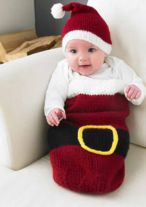 Super cute baby cocoon in a Santa inspired design. Knitted in dark red yarn with a white top. There is a black belt and gold buckle around the centre of the cocoon. The baby is wearing a white banded, dark red Santa hat with white pom pom