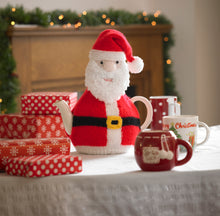 Load image into Gallery viewer, Image of a festive tea cosy displayed on a festive table with gift boxes. This Father Christmas tea cosy has a furry beard and traditional red and white hat and coat. He is finished off with a black belt and gold buckle
