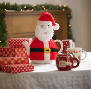 Image of a festive tea cosy displayed on a festive table with gift boxes. This Father Christmas tea cosy has a furry beard and traditional red and white hat and coat. He is finished off with a black belt and gold buckle