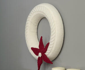 A simple white knitted wreath with a cable twist running around the front. A five pointed poinsettia is knitted in red and attached to the bottom front with a white bobble at the centre