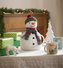 Load image into Gallery viewer, Image of festive tea cosy displayed on a festive table with gift boxes. The snowman is knitted in a textured yarn to give a snow effect. It is completed with a green, blue and red bobble hat and scarf. Down the front are 3 black buttons
