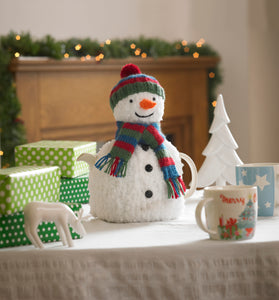 Image of festive tea cosy displayed on a festive table with gift boxes. The snowman is knitted in a textured yarn to give a snow effect. It is completed with a green, blue and red bobble hat and scarf. Down the front are 3 black buttons