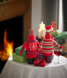 2 wine bottle jumpers sitting on a festive decorated table. One is red with a Fair Isle design - white snowflake and green and blue details. The other is red, white and green stripes with red bands and neck, topped with a white brimmed, red Santa hat