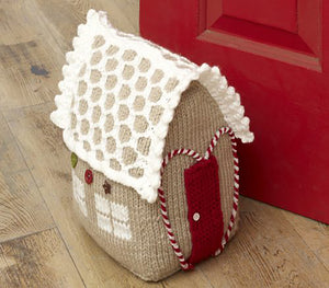A knitted gingerbread doorstop. Knitted in light brown yarn. On the side are 2 windows each made of 4 white panes of 'glass'. The red door is outlined with a red and white twisted yarn in the shape of a heart. The white latticed roof has a picot edge