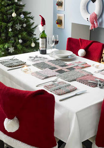 Photo of a Christmas table set for dinner. The Santa chair cover is shown along with hand knitted coasters and a table runner. There is a fun gonk or Xmas gnome wine bottle topper and a snowman wreath