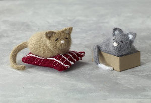 2 hand knitted kittens. They are tiny and both lying asleep. The light brown one is lying on a dark red cushion with white checks. The grey one has white ears and tail tip and is lying in a box. Both have embroidered face features