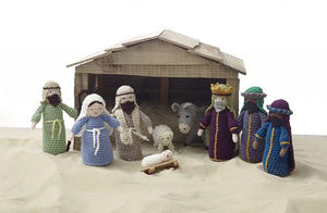 A crocheted nativity set picture on sand in front of a wooden stable. Joseph, Mary, babyJesus, donkey, sheep, shepherd and the 3 wise men. All wear robes and head dresses. One wise man has a crown and all the men have beards