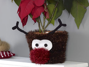 A fun Rudolph the red nosed reindeer plant pot knitted in dark brown tinsel yarn. His dark brown antlers are knitted in dark brown DK yarn. His eyes are a piece of white felt with black buttons. His nose is a red tinsel pom pom