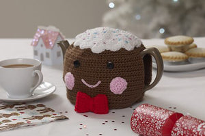 Fun Christmas pudding tea cosy. The main pudding is crocheted in brown yarn with black button eyes and embroidered pink mouth and cheeks. The top is white with coloured sprinkles and the finishing touch is a red bow tie on the front at the bottom