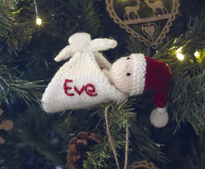 A personalised baby Christmas tree ornament. A small baby with a dark red Santa hat lies in a white shawl-shawl like hammock. The hat is trimmed in white with a white bobble. On the side of the shawl is the baby's name. Perfect for a first Xmas gift
