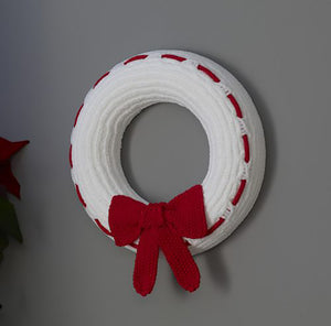 A simple wreath knitted in white chenille type yarn. A red cord is sewn in and out around the front edge and it is finished with a red seed stitch bow attached to the bottom
